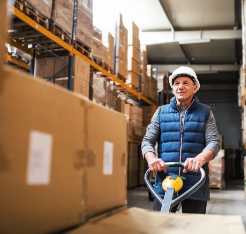 Senior male warehouse worker pulling a pallet truck with boxes.